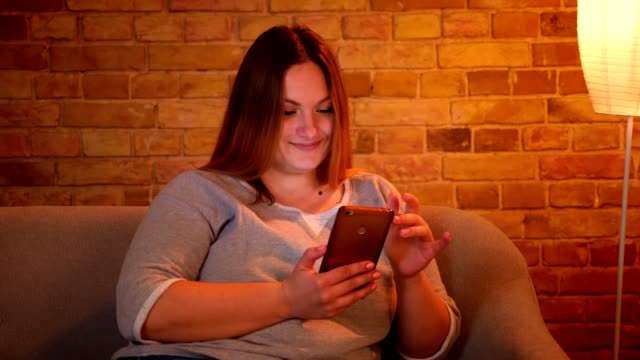 Portrait-of-relaxed-plus-size-model-fixing-her-hair-holding-a-smartphone-in-cozy-home-atmosphere.