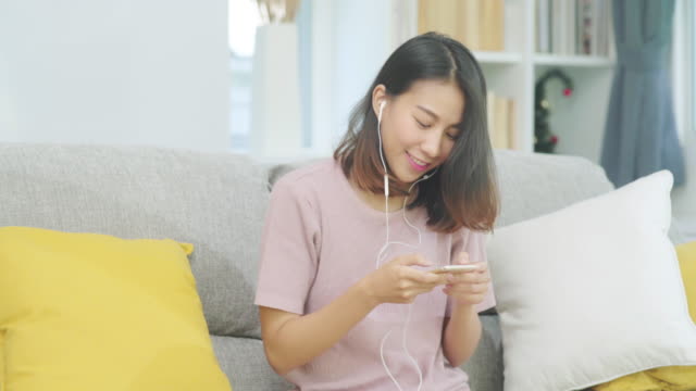 Asian-woman-listening-music-and-using-smart-phone,-female-using-relax-time-lying-on-home-sofa-in-living-room-at-home.-Happy-female-listening-music-with-headphones-concept.