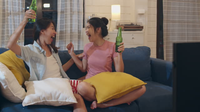 Lesbian-lgbt-women-couple-party-at-home,-Asian-female-drinking-beer-watching-TV-cheer-soccer-funny-moment-together-on-sofa-in-living-room-in-night.-Young-lover-football-fan,-celebrate-holiday-concept.