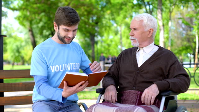 Social-worker-reading-book-for-disabled-pensioner,-leisure-in-park,-volunteering