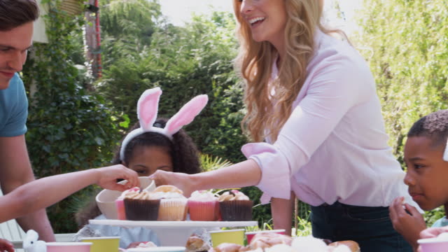 Group-of-children-wearing-bunny-ears-sitting-at-table-outdoors-enjoying-Easter-party-with-parents---shot-in-slow-motion