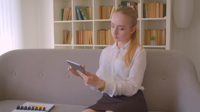 Nahaufnahme-Porträt-der-jungen-hübschen-kaukasischen-blonden-Studentin-mit-dem-Tablet-sitzend-auf-der-Couch-drinnen-in-der-Wohnung