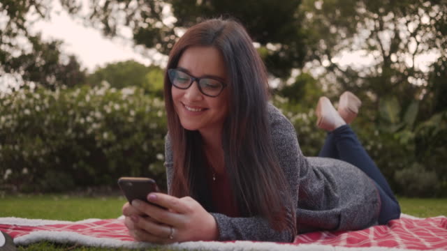 Porträt-einer-entspannt-lächelnden-jungen-Frau,-die-mit-dem-Smartphone-auf-einer-Decke-im-Park-liegt