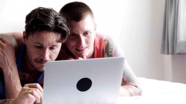 Gay-couple-in-bed-using-laptop-computer.-Focus-on-keying.