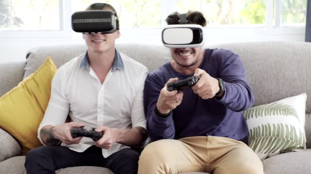 Gay-couple-relaxing-on-couch-playing-virtual-reality-games.-Exciting-mood.-Try-to-compete-together.