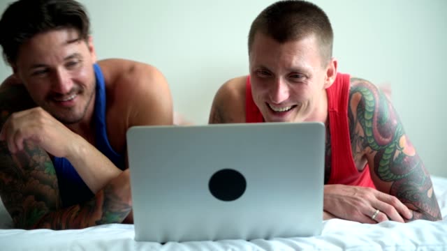 Gay-couple-in-bed-using-laptop-computer.-Pad-in-the-back.