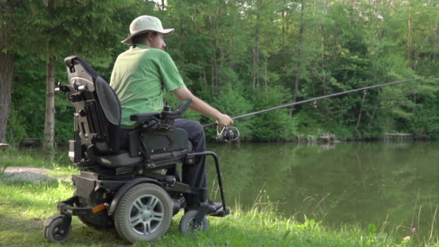 Detrás-de-la-espalda-cámara-lenta-de-pescador-discapacitado-en-una-silla-de-ruedas-eléctrica-pescando-en-el-hermoso-lago-cerca-del-bosque-y-la-montaña-en-la-parte-posterior
