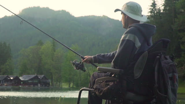 Zeitlupe-von-behinderten-Fischer-in-einem-elektrischen-Rollstuhl-Angeln-in-schönen-See-in-der-Nähe-von-Wald-und-Berg-im-Rücken,-im-Sonnenuntergang,-Sommer