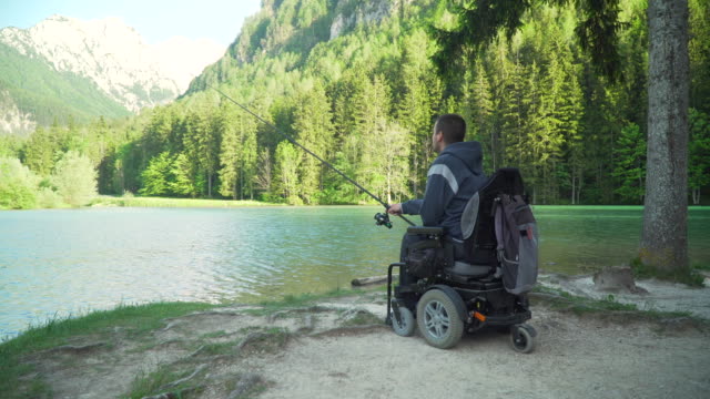 4k-resolution-of-handicapped-fisherman-in-a-electric-wheelchair-fishing-in-beautiful-lake-near-forest-and-mountain-in-the-back