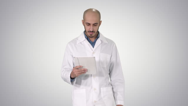 Mature-male-doctor-holding-digital-tablet-using-it-and-walking-on-gradient-background
