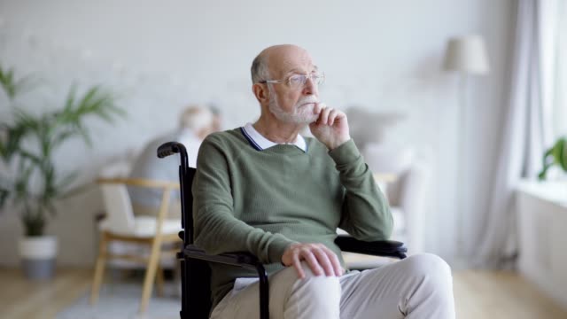 Tracking-medium-shot-of-disabled-elderly-man-in-eyeglasses-sitting-in-wheelchair-and-thinking-dreamily,-then-looking-at-camera-and-smiling-happily-in-nursing-home
