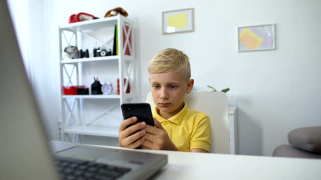 Little-male-kid-sitting-in-front-of-laptop-and-watching-video-on-smartphone,-app