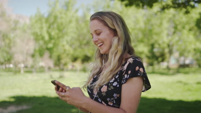 Cheerful-attractive-blonde-young-woman-standing-in-the-park-with-earphones-in-her-ears-using-mobile-phone-smiling-and-happy