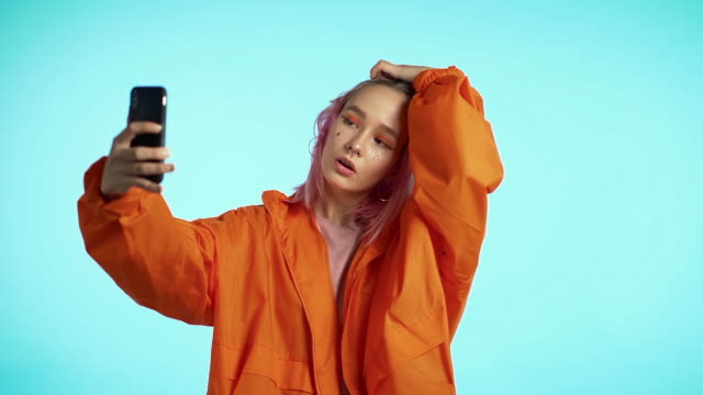 Mädchen-mit-rosa-Haaren-und-außergewöhnlichem-Aussehen-machen-Selfie-auf-blauem-Hintergrund.-Mit-moderner-Technologie---Smartphone,-soziale-Netzwerke