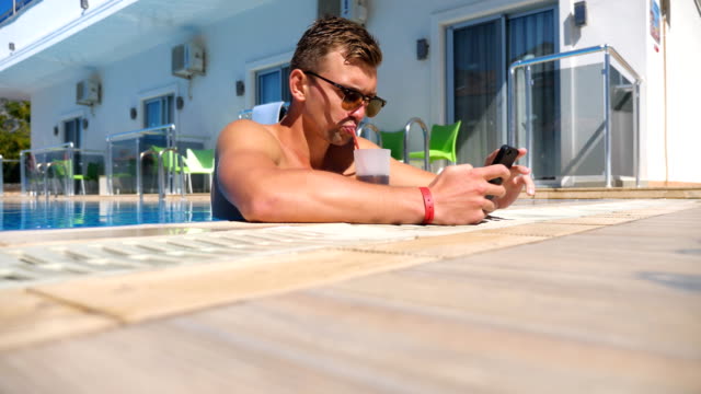 Man-using-phone-at-pool-and-refreshing-cold-cocktail.-Guy-drink-beverage-browsing-internet-page-or-communicating-with-somebody.-Young-man-enjoying-recreation-on-resort-during-summer-vacation.-Low-view