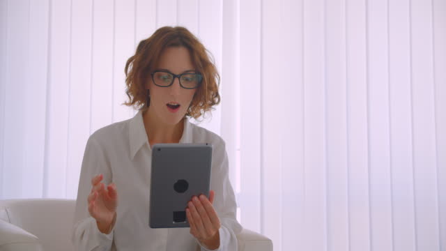 Nahaufnahme-Porträt-der-erwachsenen-Rotschopf-kaukasische-Geschäftsfrau-in-Brille-mit-einem-Videoanruf-auf-dem-Tablet-winken-hallo-sitzend-im-Sessel-im-weißen-Büro