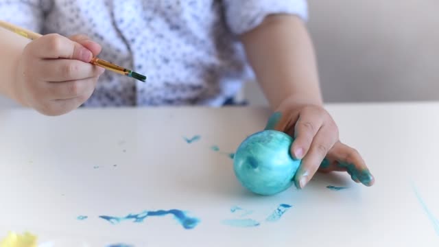 The-hands-of-a-litlle-child-with-a-brush-paint-an-Easter-egg-in-blue-color-on-the-table.