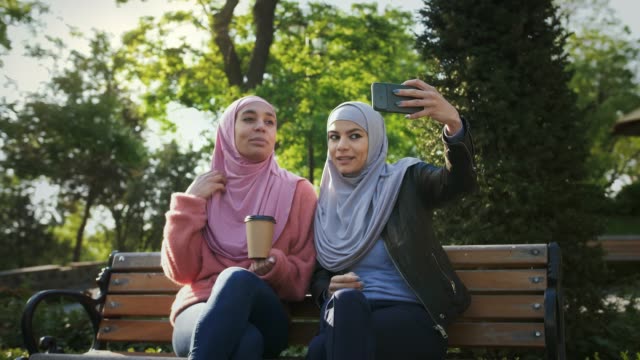 Dos-señoras-musulmanas-en-hiyabs-coloridos.-Sonríen,-se-toman-selfies-en-el-teléfono-inteligente-y-disfrutan-de-un-café-mientras-se-sientan-en-el-banco-en-el-parque.-"Lose-up"