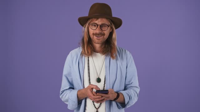 Long-haired-fellow-in-casual-clothes,-glasses,-hat.-Smiling,-using-smartphone,-looking-surprised-and-saying-wow.-Posing-on-purple-background.-Close-up