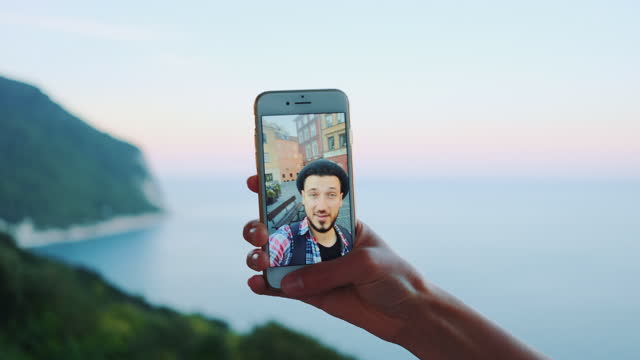 Close-up-of-hand-holding-smartphone-during-video-call-with-man-in-front-of-the-sea