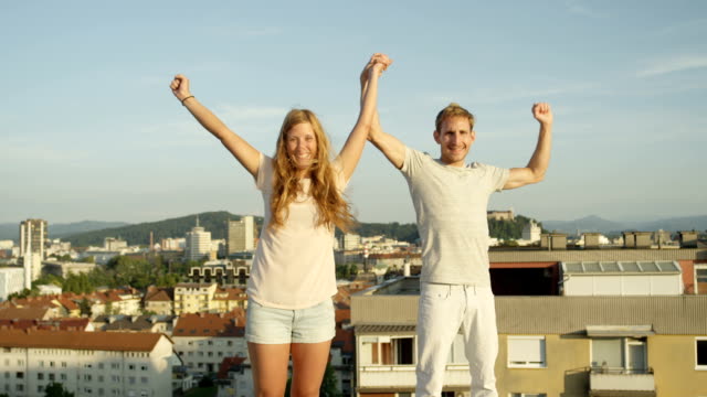 CLOSE-UP:-Happy-boy-and-smiling-girl-standing-on-rooftop-raising-hands-in-sky