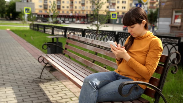 Young-woman-using-telephone-sitting-on-the-bench-near-the-road