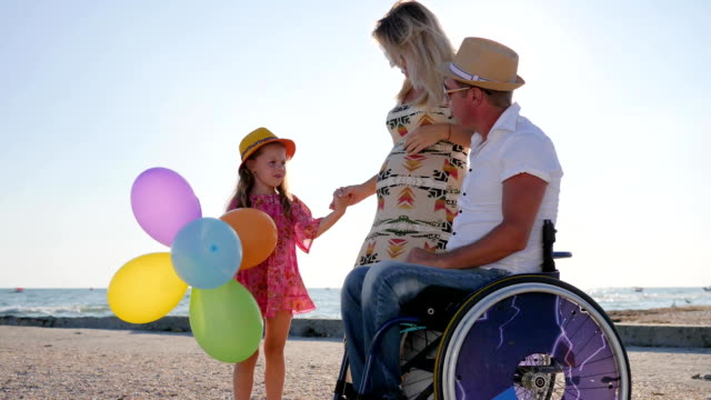 parents-tell-of-maternity,-disabled-person-in-wheelchair-with-family,-little-girl-listens-mother-and-father-on-wheel-chair-at-beach