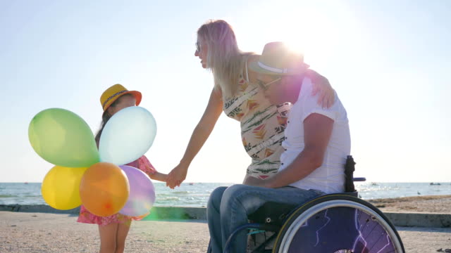 disabled-person-in-wheelchair-with-family,-motherhood,-little-girl-listens-mother-and-father-on-beach-in-summer-time,-happy-mom-and-daughter-stand-near-dad