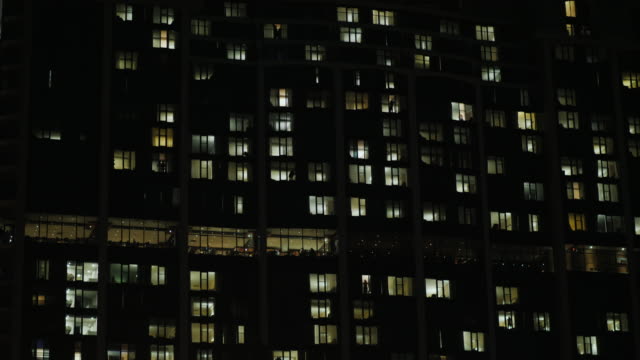 Bürogebäude-in-der-Dunkelheit.-Die-Fenster-sind-beleuchtet,-Menschen-Silhouetten-sind-sichtbar.-Schuss-zu-kippen