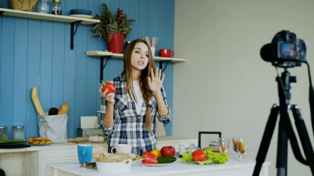 Mujer-atractiva-alegre-video-blog-sobre-comida-saludable-vegetariana-en-cámara-réflex-digital-de-grabación-en-cocina-en-casa