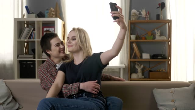 Zwei-junge,-schöne-Mädchen-auf-der-Couch-sitzen,-küssen-und-machen-Selfies,-Lesben,-LGBT,-blond,-brünett-60fps