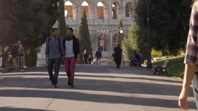 Young-happy-gay-couple-tourists-walk-in-park-road-with-trees-colosseum-in-background-in-rome-at-sunset-holding-hands-lovely-slow-motion-colle-oppio