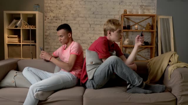Two-young-mans-are-sitting-on-the-couch-and-using-a-smartphones,-gays,-the-problem-of-society,-a-new-generation,-lgbt-lovers,-homo,-homosexuality-concept-60-fps