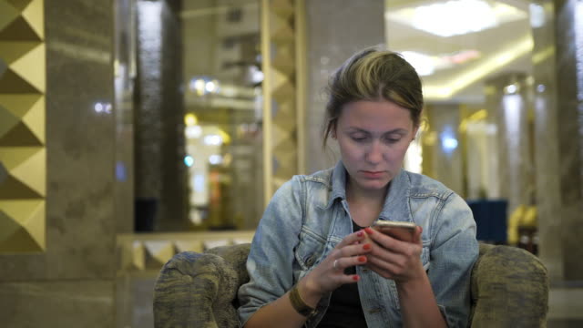 young-Caucasian-woman-in-a-jeans-jacket-drinks-juice-from-a-glass-and-uses-a-smartphone,-writes-messages-on-the-social-network.-Sits-at-the-table-in-the-hotel-restaurant.-Concept-of-healthy-fresh-food-in-business.-Close-up