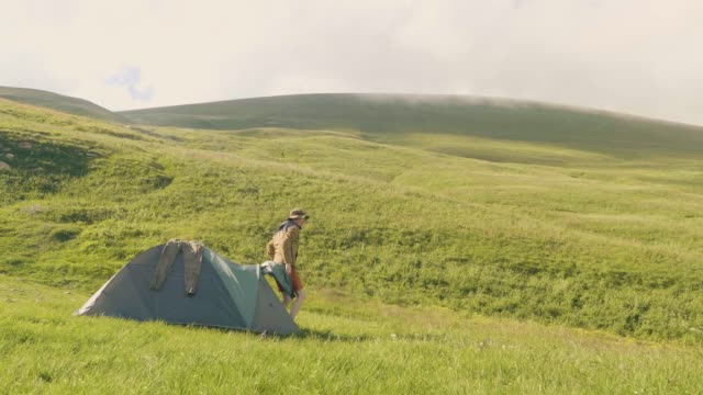 Tourist-man-came-out-of-camping-tent-on-green-meadow-and-mountain-background