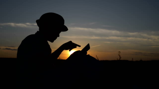 Silhouette-eines-jungen-verwendet-eine-Tablette-bei-Sonnenuntergang-im-Feld-liest-etwas-auf-dem-tablet