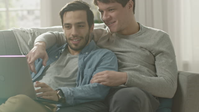 Sweet-Male-Queer-Couple-Spend-Time-at-Home.-They-are-Lying-Down-on-a-Sofa-and-Use-the-Laptop.-They-Browse-Online.-Partner's-Hand-is-Around-His-Lover.-They-Smile-and-Laugh.-Room-Has-Modern-Interior.