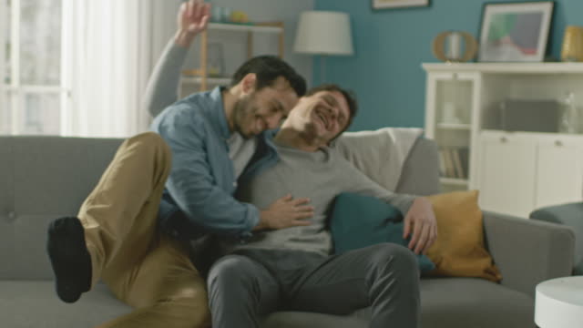 Slow-Motion-of-a-Male-Gay-Couple-Fulling-Around-on-a-Sofa-at-Home.-Boyfriend-Runs-and-Jumps-into-Hands-of-His-Partner.-They-Hug.-They-are-Happy-and-Laughing.-They-are-Casually-Dressed-and-Room-Has-Modern-Interior.