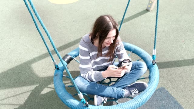 Beautiful-brunette-girl-is-swinging-on-empty-webbed-swing,-typing-a-message-on-mobile-phone.
