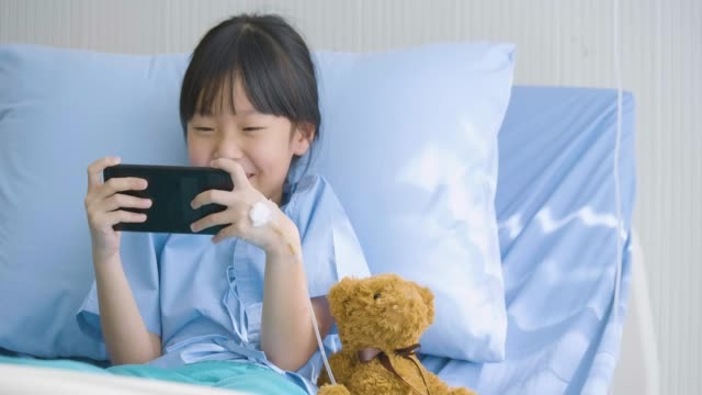 Niña-linda-acostado-en-cama-en-el-hospital,-viendo-dibujos-animados-divertidos,-películas-en-smartphone.-enfermedad-y-tratamiento.