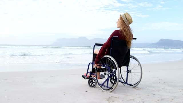 Vista-posterior-de-los-jóvenes-con-discapacidad-mujer-caucásica-con-sombrero-sentado-en-silla-de-ruedas-en-playa-4k
