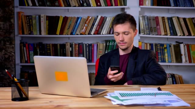 Atractivo-estudiante-masculino-sentado-en-frente-de-la-computadora-portátil-y-la-navegación-en-el-teléfono-en-la-biblioteca-en-el-interior