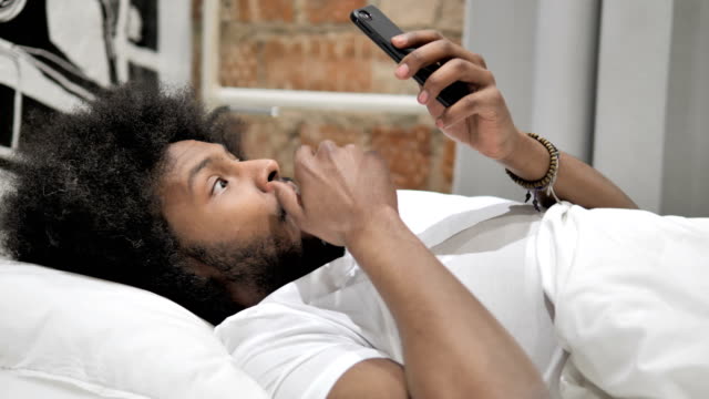 Erschütterner-afrikanischer-Mann-mit-Smartphone-in-Bett