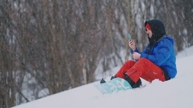 Ein-männlicher-Snowboarder,-der-auf-dem-Schnee-sitzt,-fotografiert-am-Telefon-eine-schöne-Resort-Landschaft-für-soziale-Netzwerke.-Blogger-im-Resort.-Schreiben-Sie-Textnachrichten-an-Freunde-mit-Ihrem-Smartphone