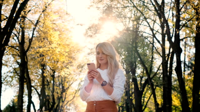 Stilvoll-junge-niedlich-lächelnde-Mädchen-mit-modernen-Smartphones-beim-Gehen-in-der-Stadt-Park.-Lady-tippen-Textnachricht-auf-Handy-scrollt-durch-soziale-Medien,-Nachrichten-auf-App,-bei-Sonnenuntergang