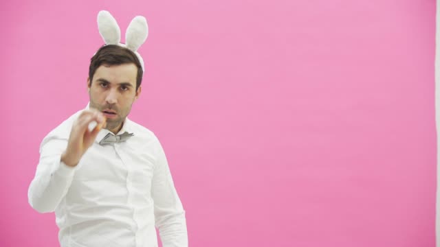 Young-man-standing-on-a-pink-background.-During-this-he-looks-at-the-camera-and-smokes-a-carrot.-After-putting-a-hand-on-the-arm.-The-guy-wears-a-bunny-ear.