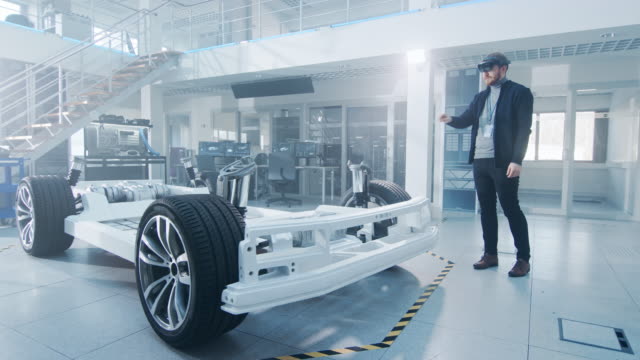Ingeniero-automotriz-trabajando-en-la-plataforma-de-chasis-de-coche-eléctrico,-utilizando-cascos-de-realidad-aumentada.-En-el-laboratorio-de-innovación,-el-marco-del-vehículo-conceptual-incluye-ruedas,-suspensión,-motor-y-batería.