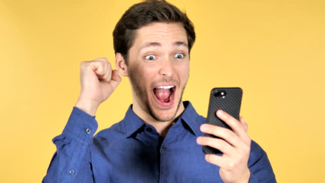 Casual-Young-Man-Celebrating-Success-while-Using-Smartphone-on-Yellow-Background