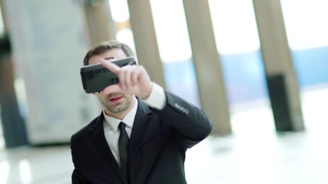 Tilt-down-shaky-shot-of-modern-businessman-in-suit-wearing-VR-headset-to-visualize-data-and-exploring-it-standing-in-office-hall