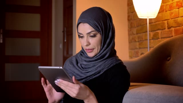 Closeup-portrait-of-adult-attractive-muslim-female-in-hijab-surfing-web-on-the-tablet-while-sitting-on-the-floor-in-doorway-in-a-cozy-apartment